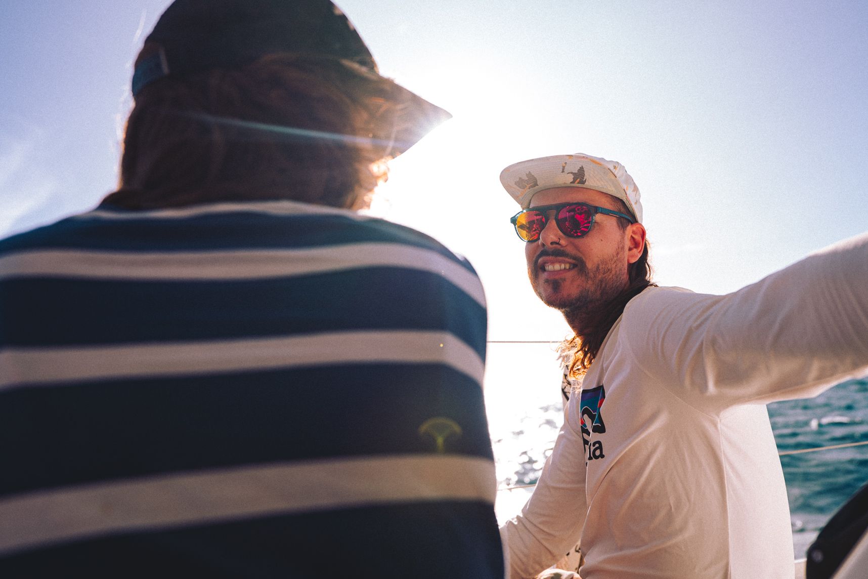 Posé sur le côté du catamaran, deux personnes discutent. Le soleil passe à travers les cheveux. Les lunettes orange reflètent la deuxième personne