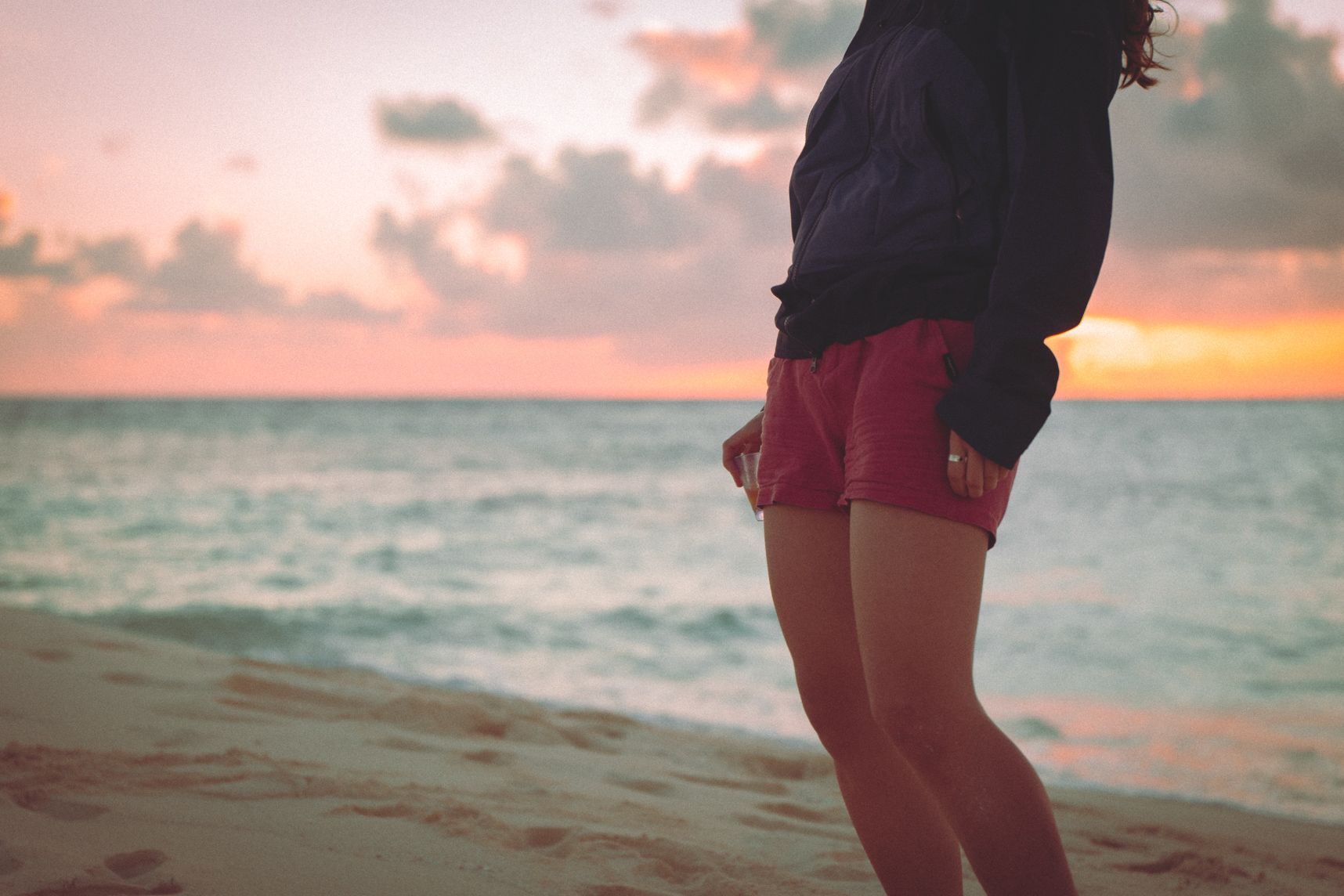 Sur une plage une personne tient un verre de punch au niveau de la taille. La photo est centrée dessus, la tête et les pieds sont coupés. Derrière la mer et le coucher de soleil dans les nuages rosés