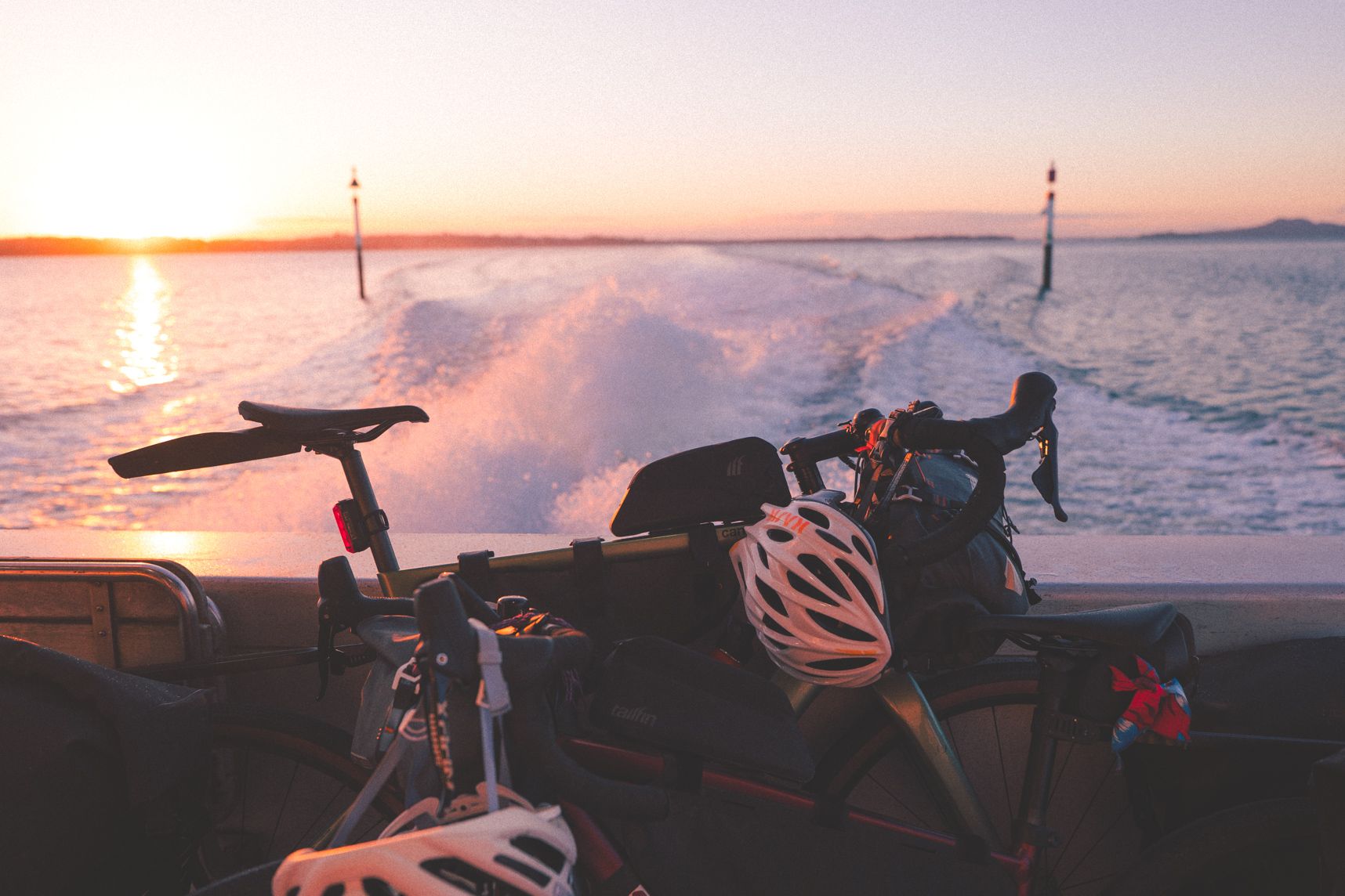 Des vélos adossés à la rambarde du bateau. Derrière les remous des moteurs dans l'eau, l'entrée du port et le coucher de soleil