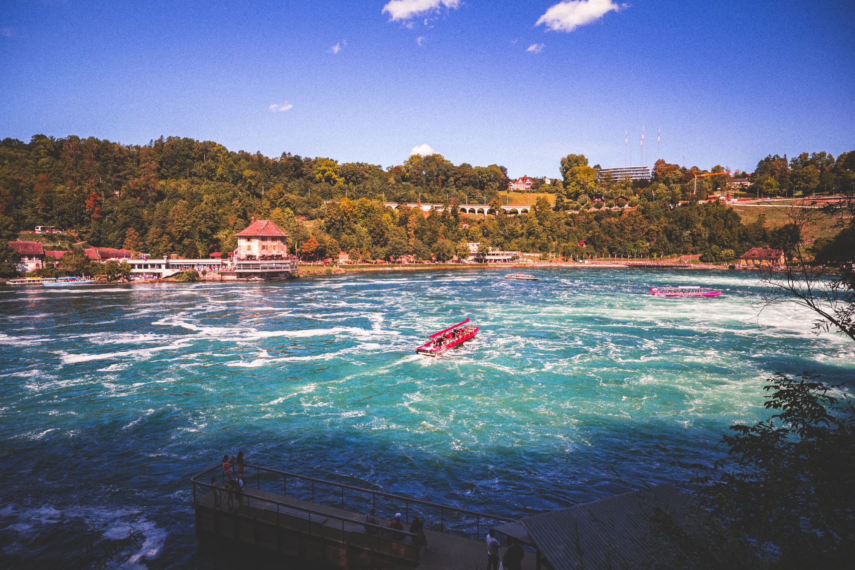 Au pied des chutes du Rhin l'eau remue. Des bateaux de touristes rouges y naviguent. L'étendue d'eau est bordée d'une petite colline boisée et bâtiments pour touriste