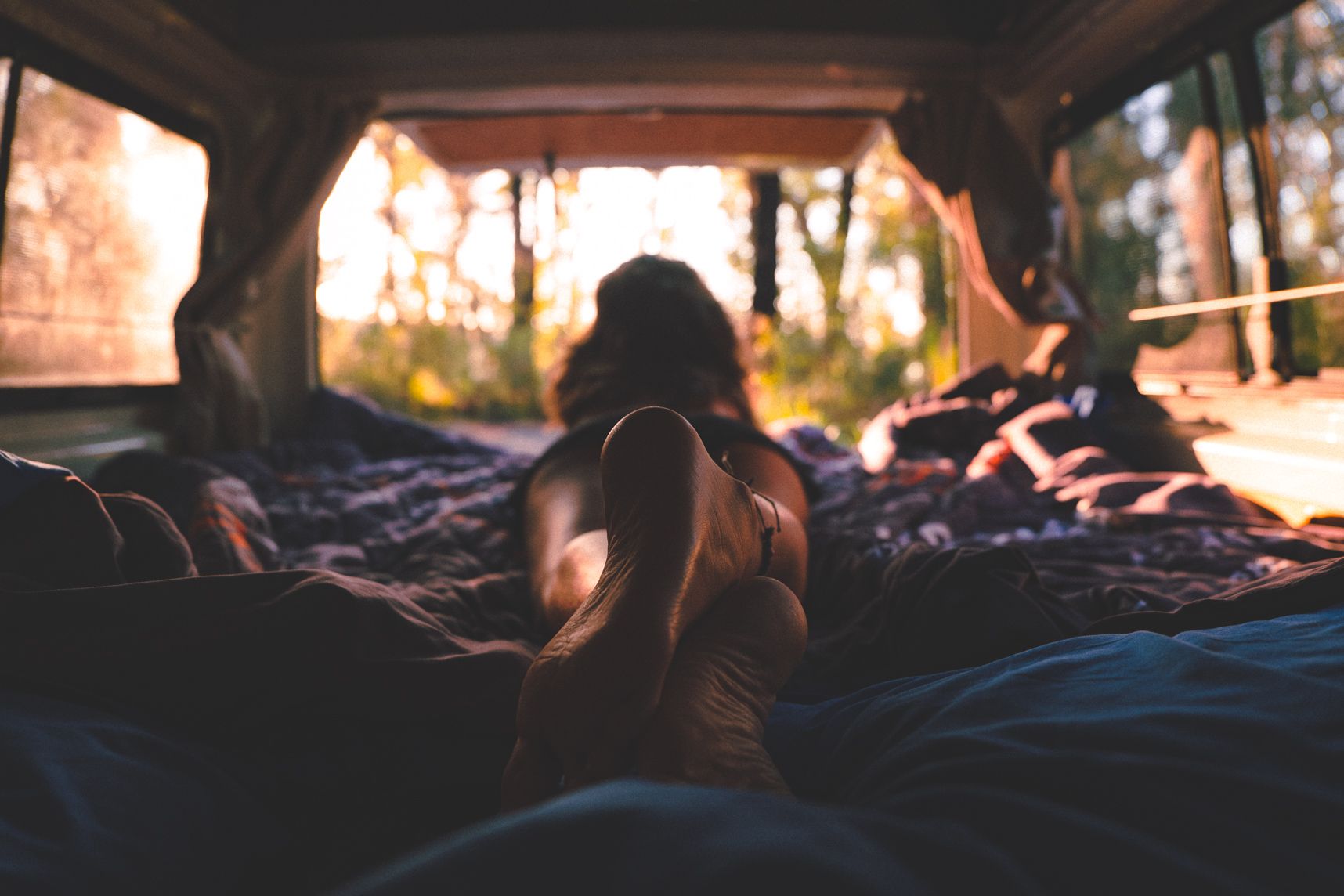 Allongée sur le ventre dans le lit du van regardant vers l'extérieur. Premier plan sur les pieds croisés, second plan l'arrière du van et le lit. Le soleil vient de se lever et illumine l'intérieur. En arrière plan la forêt