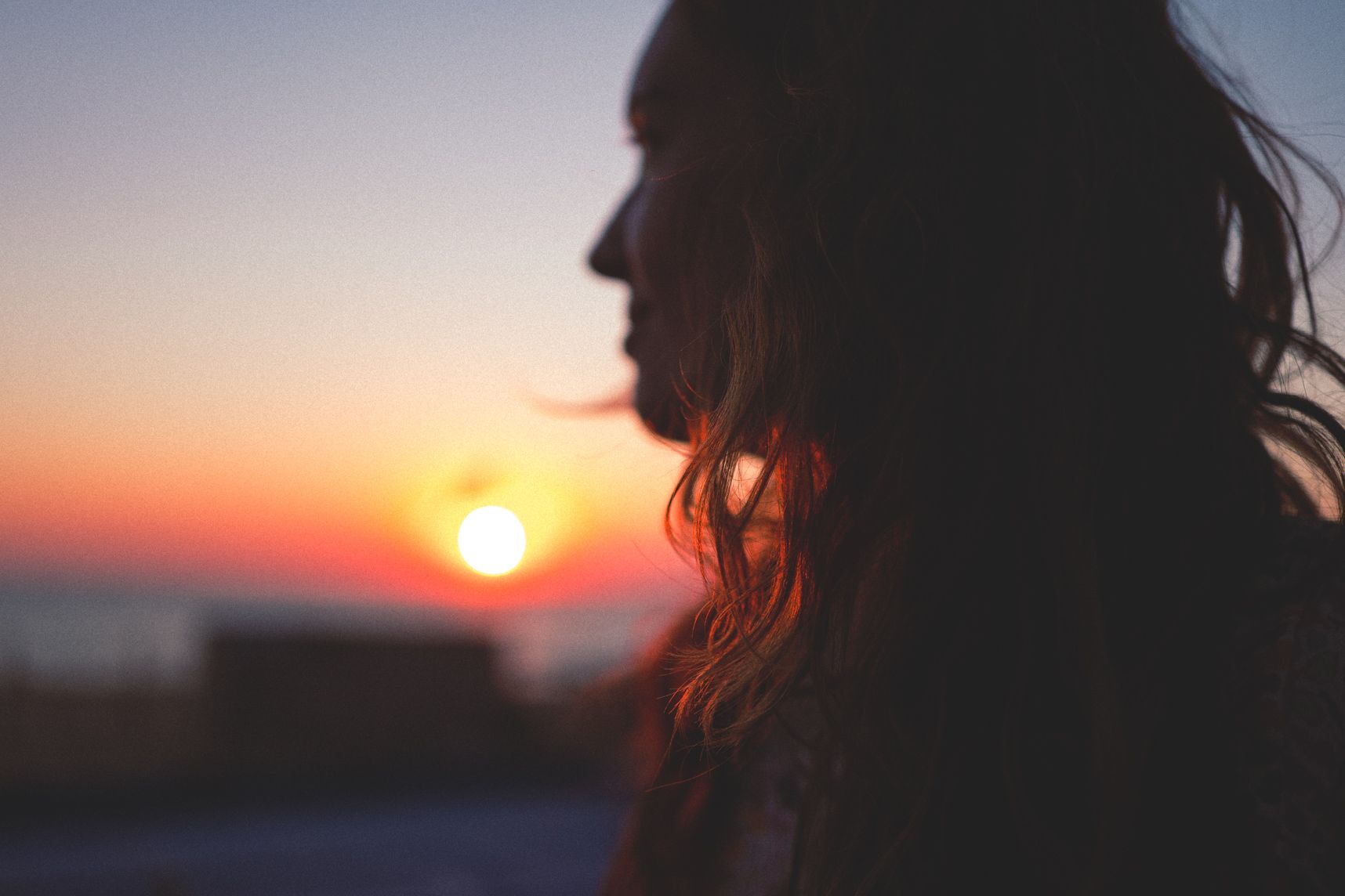 Une femme de profil avec les cheveux ondulés regarde le coucher de soleil. Le focus est sur les cheveux. Le soleil éclaire les cheveux en dessous du menton