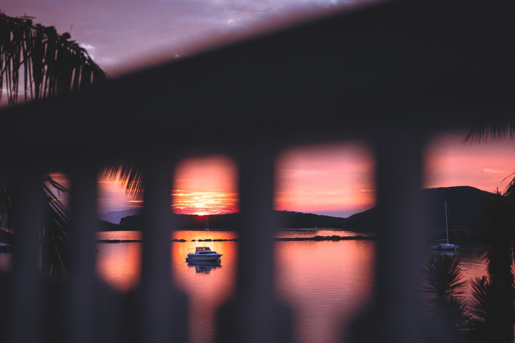 Entre la rambarde, le soleil se lève sur la baie de Ouémo. Le ciel est orangé. Une bateau est dans l'alignement du soleil