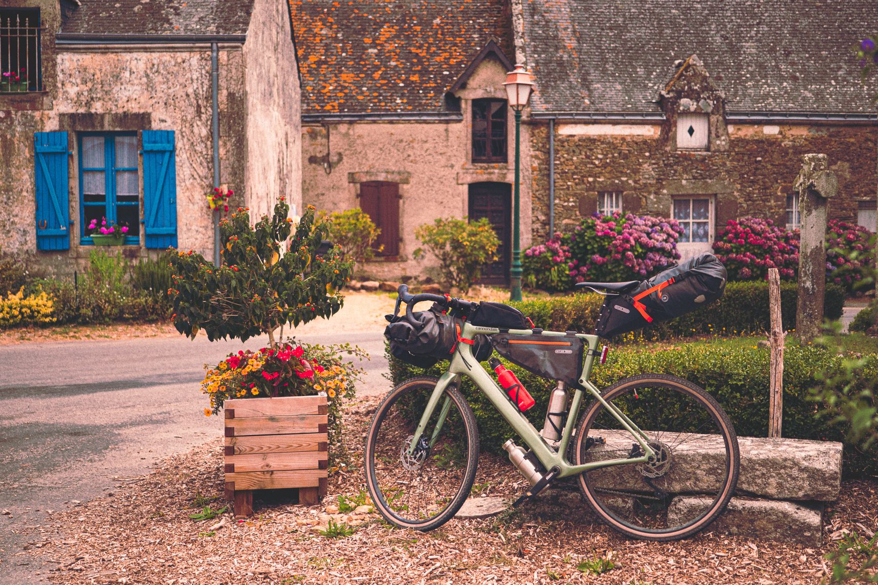 Le vélo, chargé de sacs de bikepacking, est accosté à un banc en pierre. Plein de fleurs de toutes les couleurs l'entour. En arrière plan des maisons typiques de la Bretagne