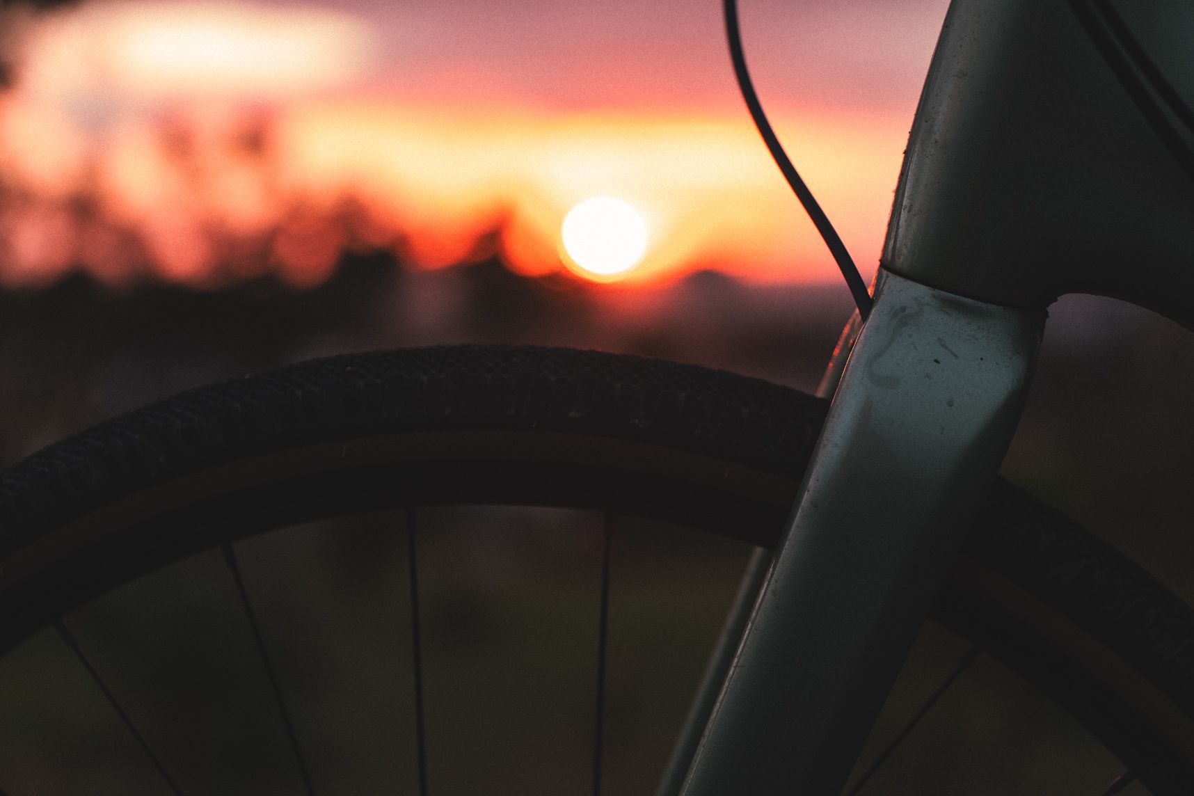 Gros plan sur le haut d'une fourche de vélo et de sa roue. Elles sont un peu sale. En arrière plan dans le flou, le coucher de soleil et ses couleurs orangées