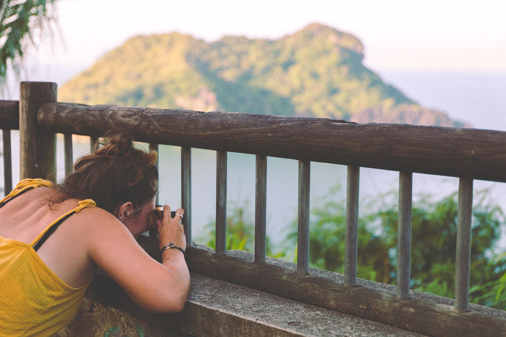 Une personne s'applique sur la cadrage du paysage avec un appareil photo jetable. Elle est entre les barreaux de la rambarde. Au loin une montagne sort de l'eau