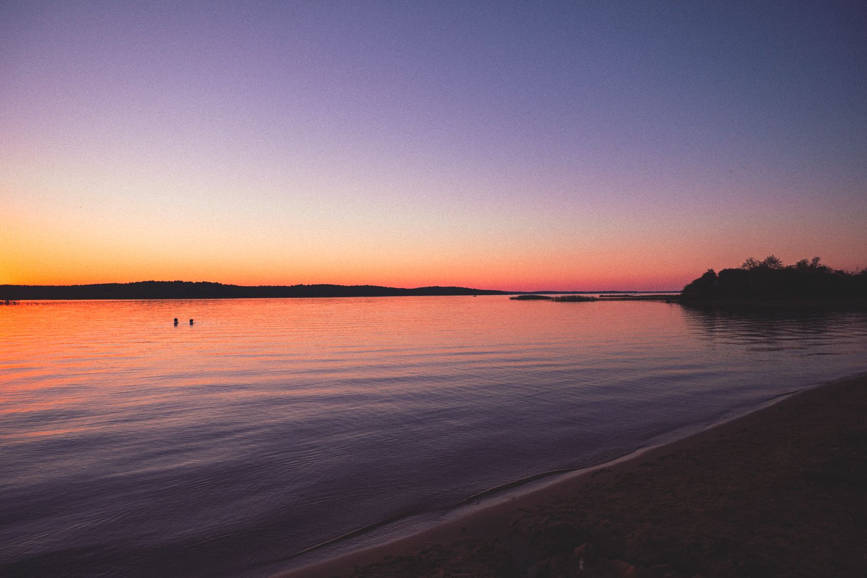 Le lac est calme et le soleil donne ses dernières lueurs. Le ciel est un dégradé orange qui reflète sur l'eau. Deux personnes discutent dans l'eau