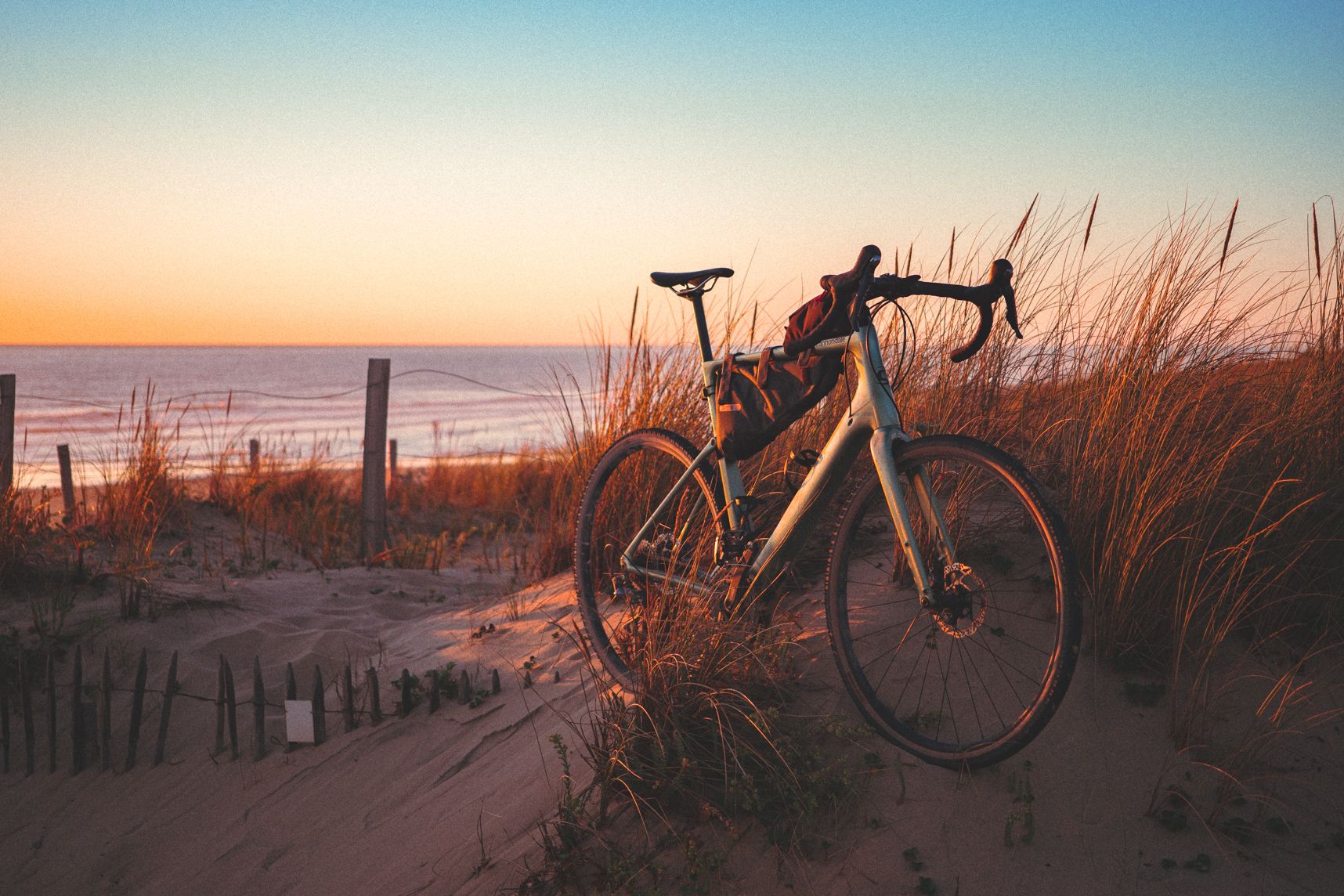 Le vélo est sur la dune entre les plantes. En arrière plan la mer et le coucher de soleil