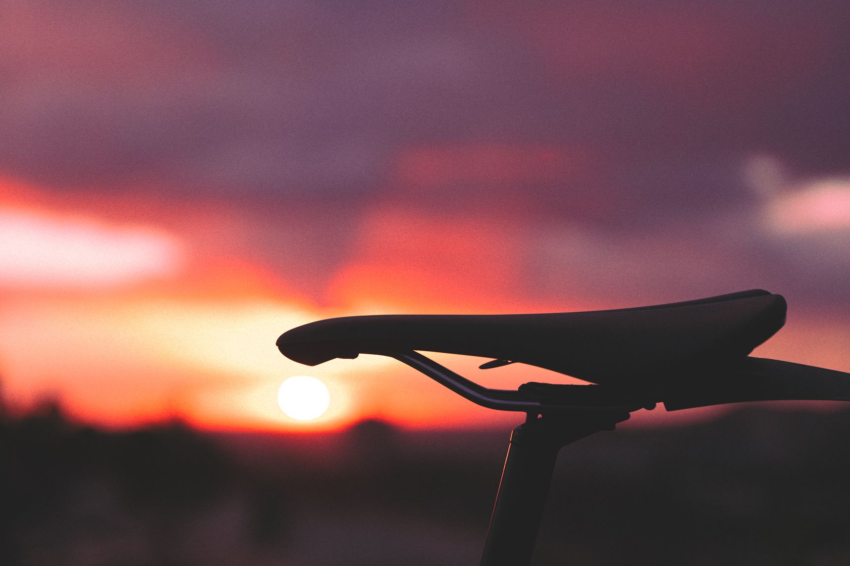 Gros plan sur une selle de vélo. Elle est profilée. En dessous du devant de la selle, le coucher de soleil et ses couleurs orangées