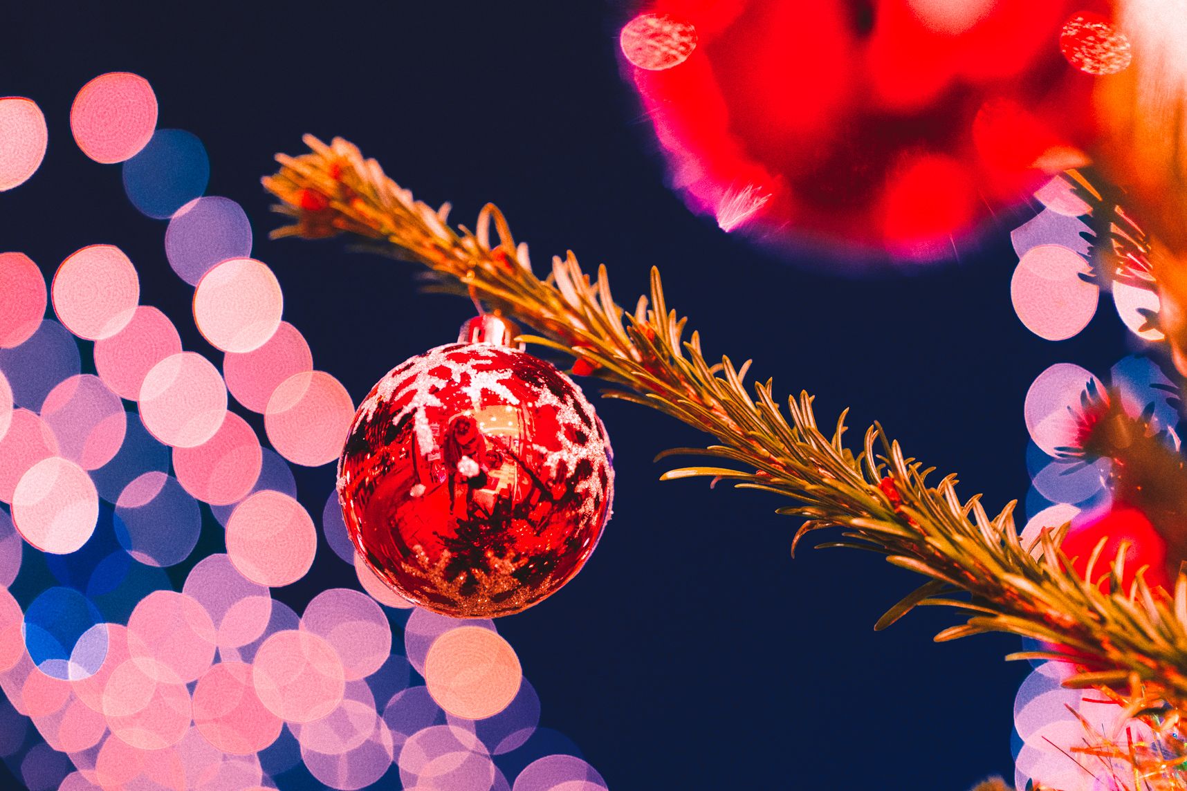 Gros plan sur une boule de Noël et sa branche de sapin. On y voit les reflets du photographe. Derrière les illuminations forment un beau bokeh