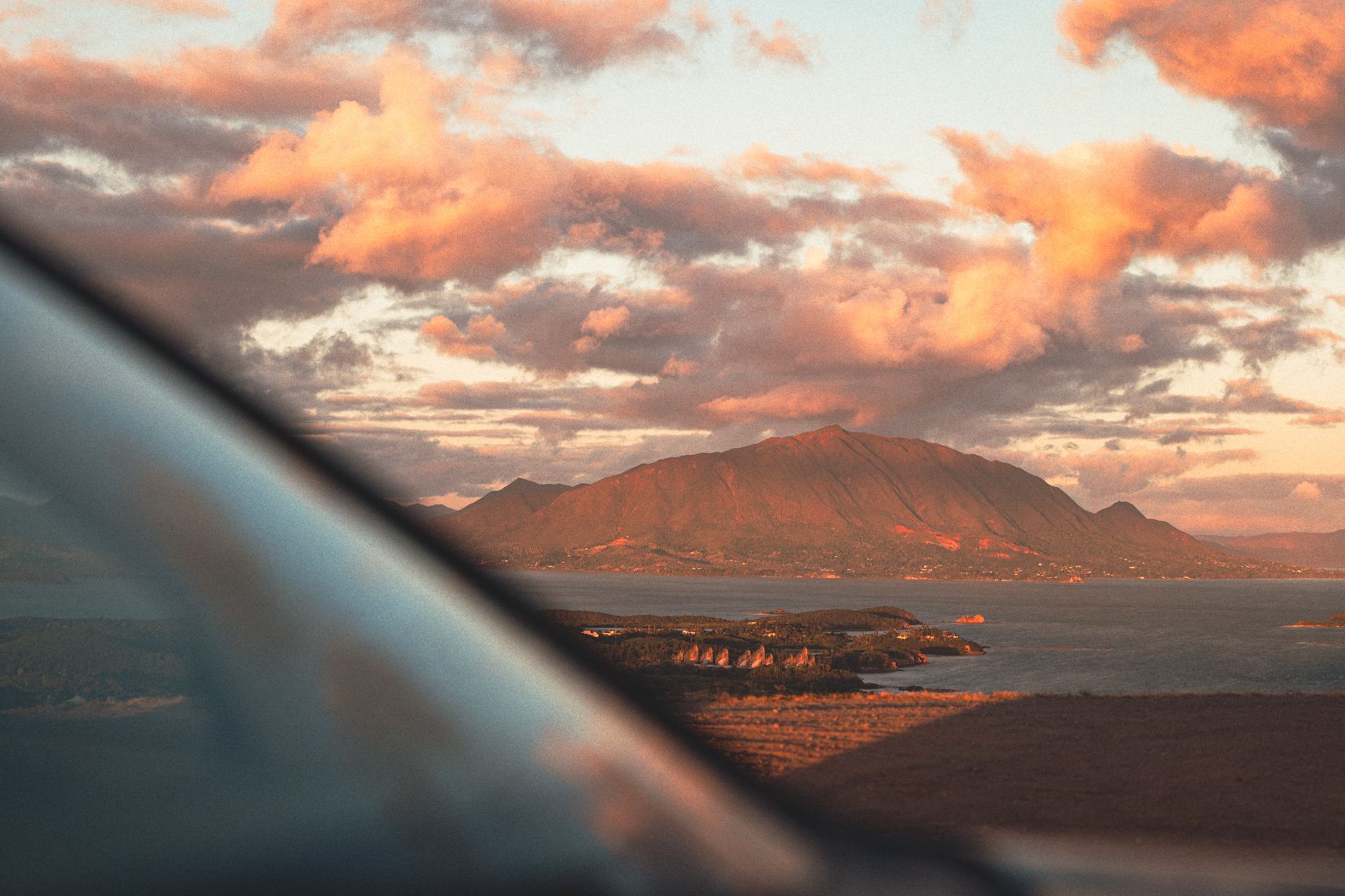 En premier plan le pare-brise d'une voiture qui reflète le ciel. Dans la continuité le centre Tjibaou et le Mont Dore baignés par le coucher de soleil