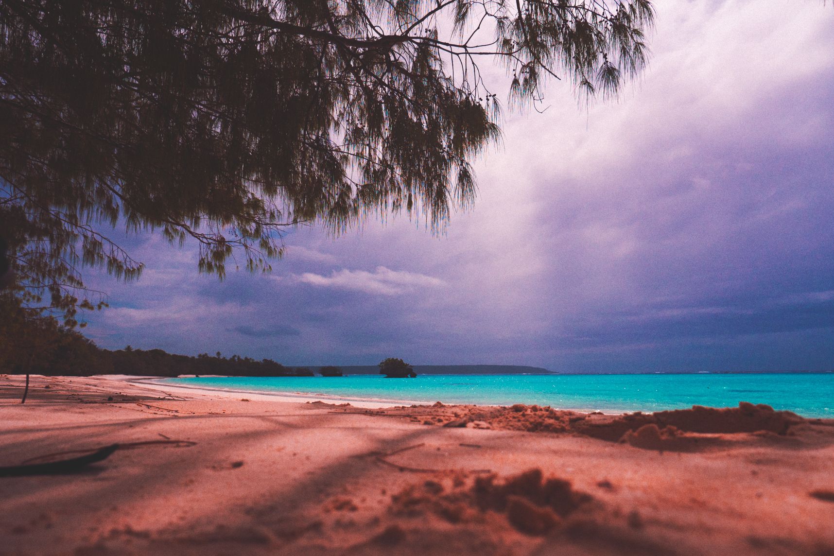 Sur la plage de Lifou le soleil parvient à passer quelques rayons à travers le ciel gris. L'eau est turquoise, l'ombre des arbres se déporte sur la plage