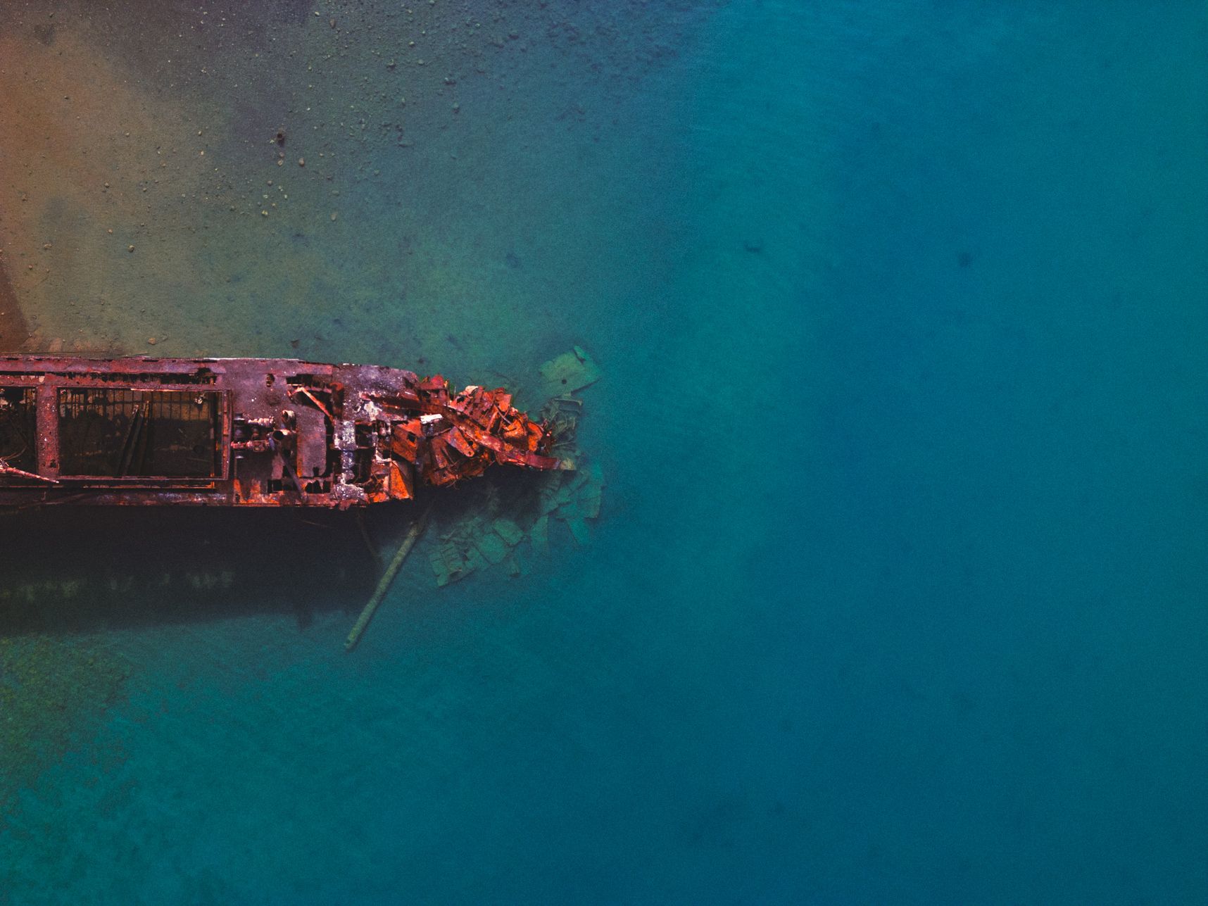 Vue du dessus de drone, l'arrière d'une épave de bateau mise dans l'eau. Des parties totalement rouillées sont hors de l'eau tandis que l'on voit quelques pièces en dessous