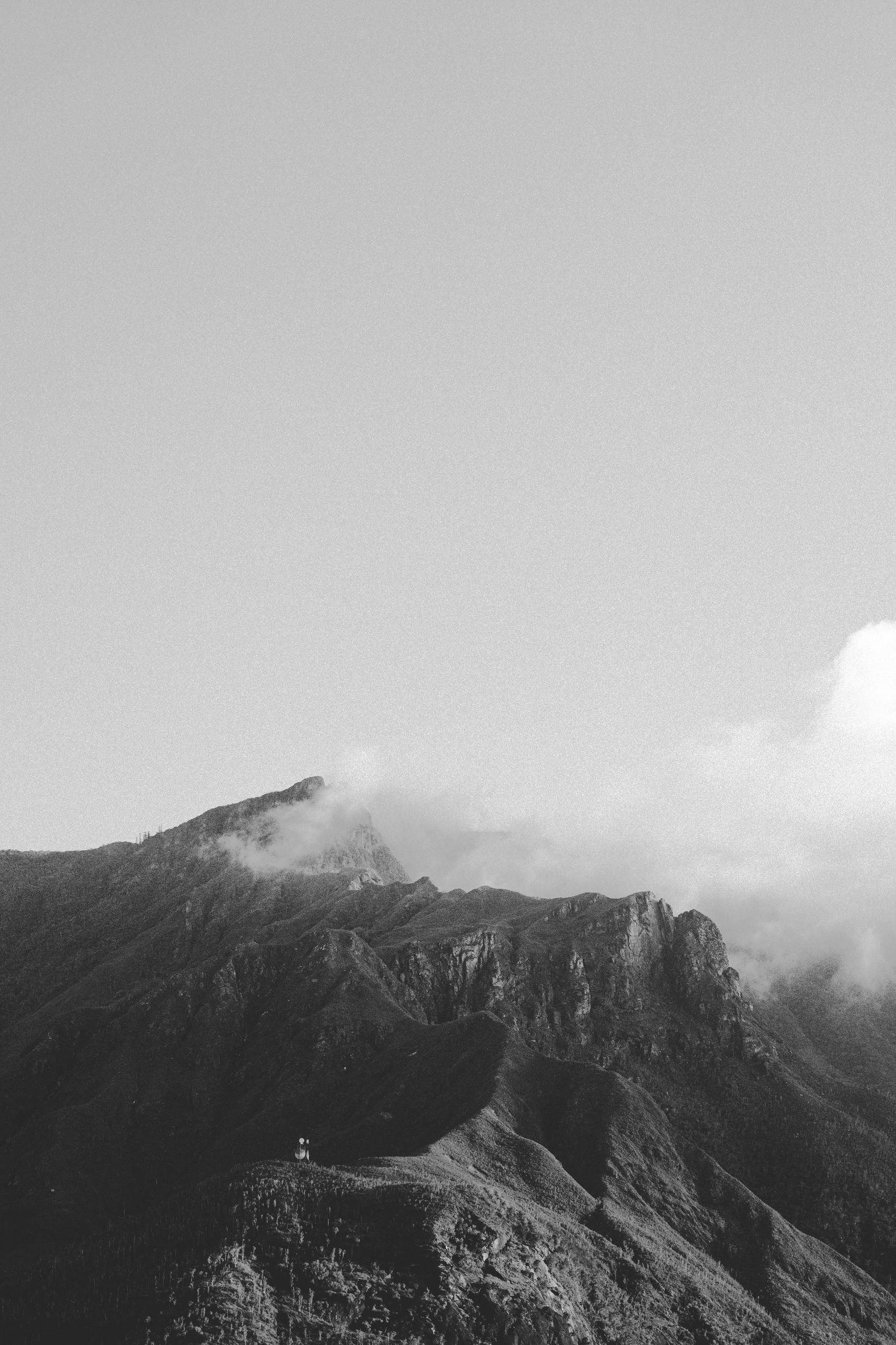 Photo en noir et blanc. La montagne vue du bas, sur la crête stagne les nuages