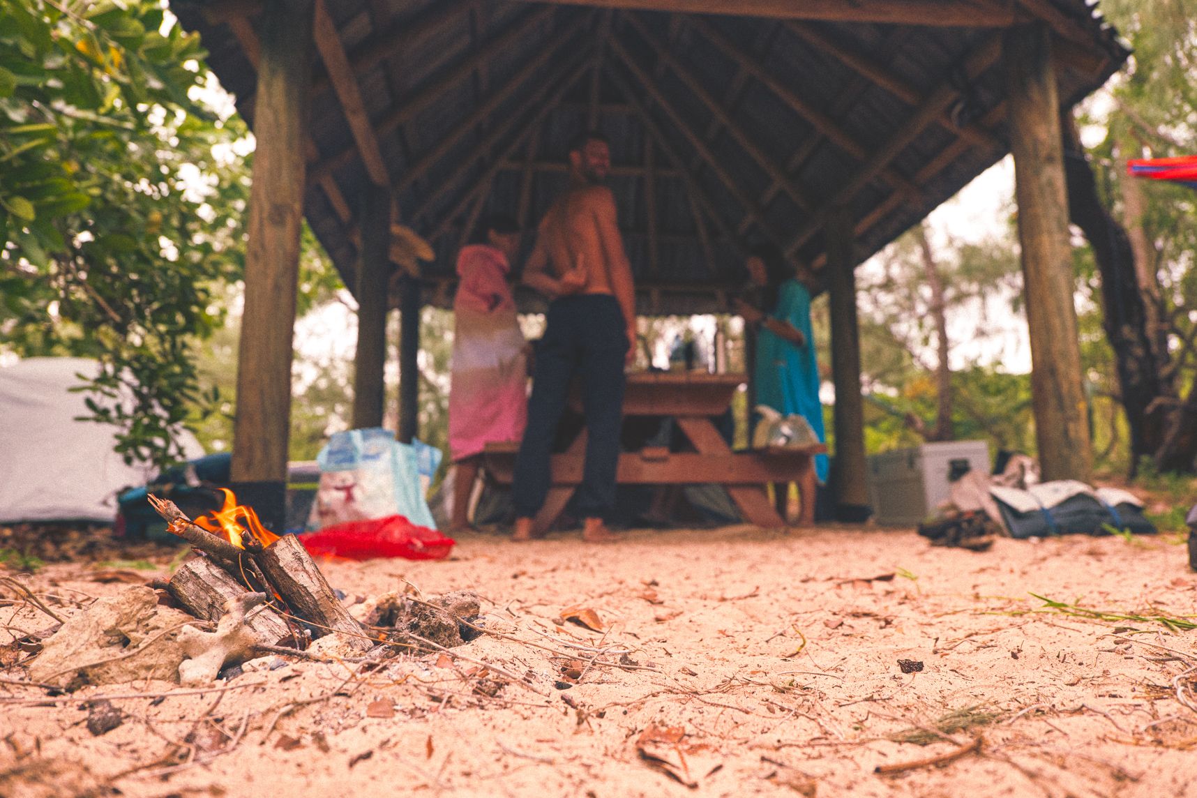 Dans le sable, des petits rondins de bois brulent. Derrière sous le faré des gens s'affairent autour de la table