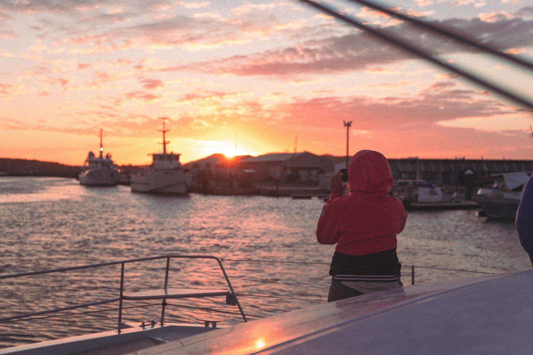 Le soleil se coucher sur le port Moselle. Sur le catamaran au premier plan, quelqu'un prend une photo. En arrière plan les bateaux et les couleurs orangées
