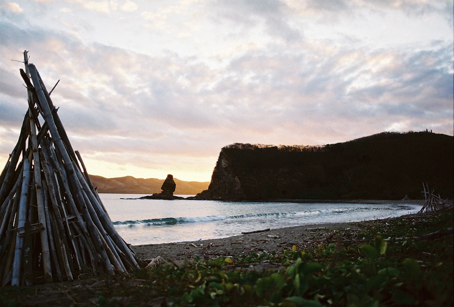Bonhomme de la Roche Percée au coucher de soleil. En premier plan un tipi en bois flotté, la mer puis derrière la falaise, le ciel nuageux qui commence à prendre des couleurs