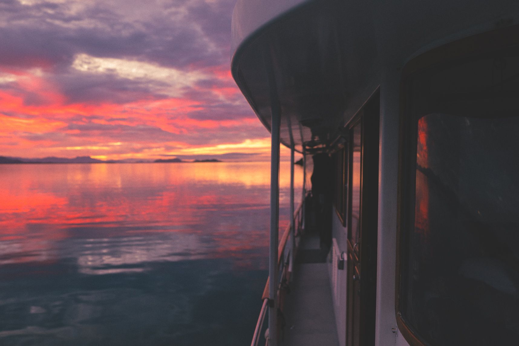 Vue du ponton du bateau, en premier plan la porte du poste de pilotage, en arrière plan le lever du soleil qui reflète sur la mer d'huile et les fenêtres