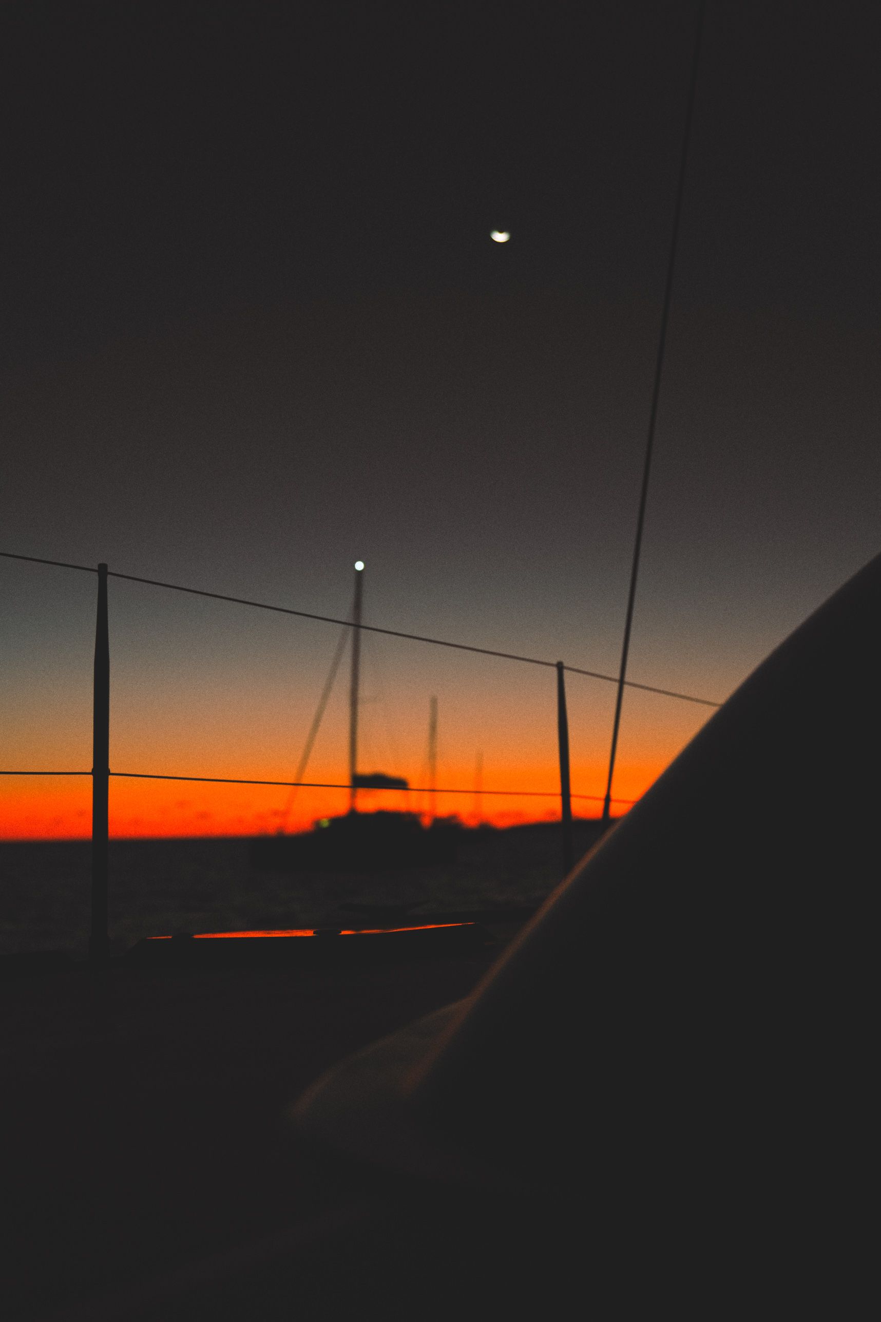 Dernières lueurs du soleil. En premier plan la rambarde d'un catamaran. En second plan dans le flou, 3 mats de catamaran alignés. Au dessus la lune.