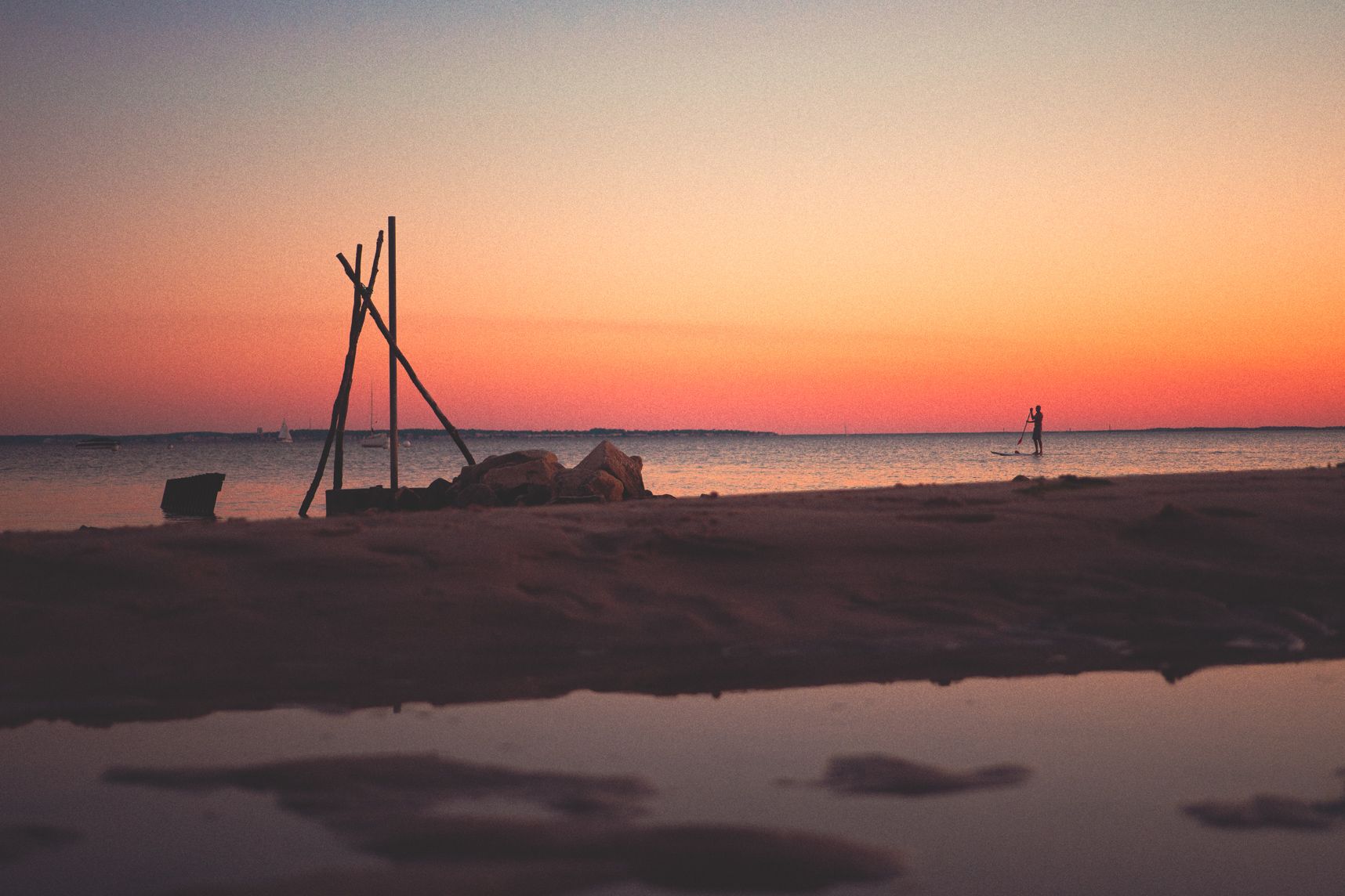 Sur le bassin d'Arcachon, quelqu'un au loin fait du paddle. C'est le coucher de soleil, le ciel est orangé. Au premier plan la plage et le reflet de flaques
