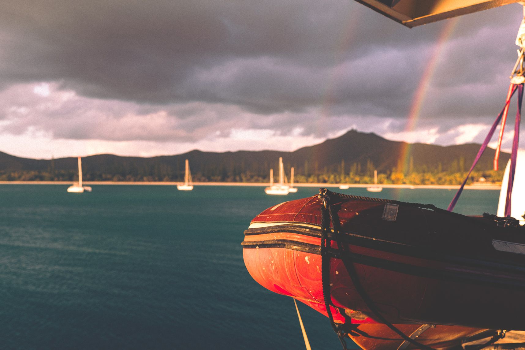 Face avant d'un zodiac de secours sur le Betico, le soleil est couchant. Les bateaux de la baie de Kuto sont éclairés, derrière 2 arc en ciel et la montagne sous les nuages