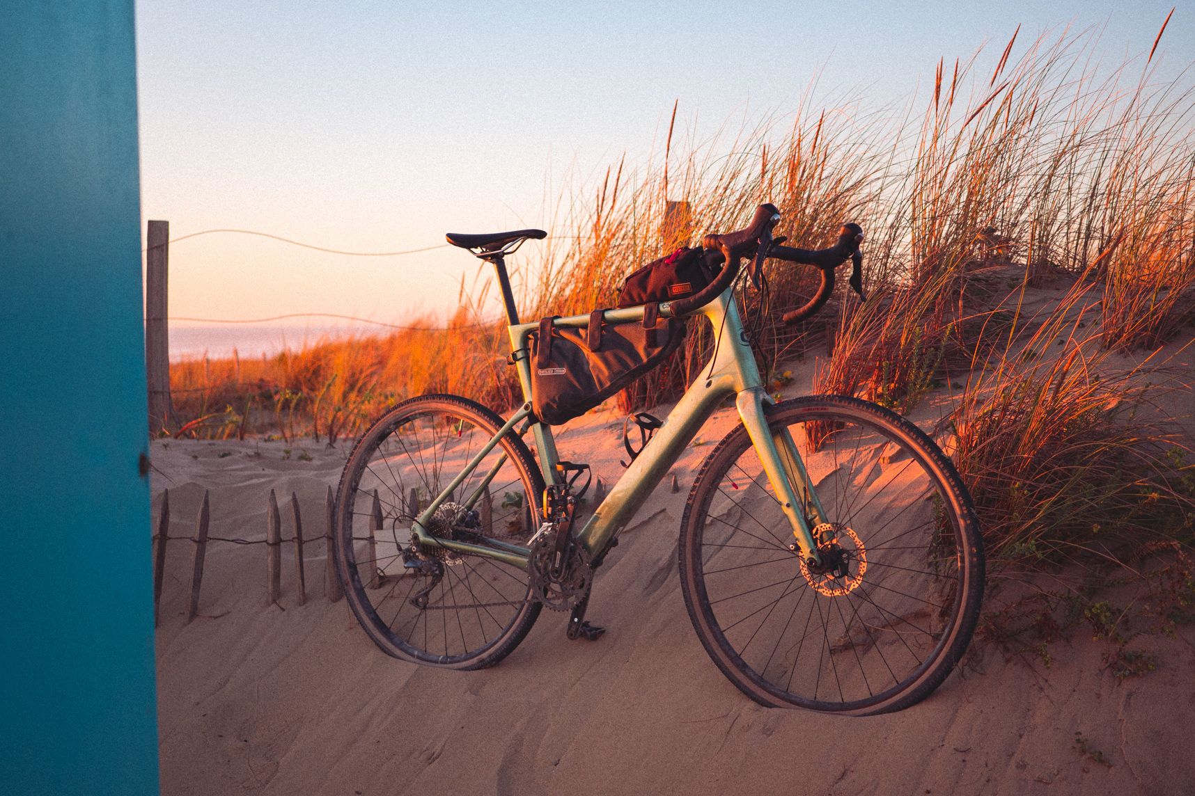 Le vélo est posé sur la dune. Une moitié est dans l'ombre, l'autre éclairée par le coucher de soleil. Des grandes herbes en arrière plan