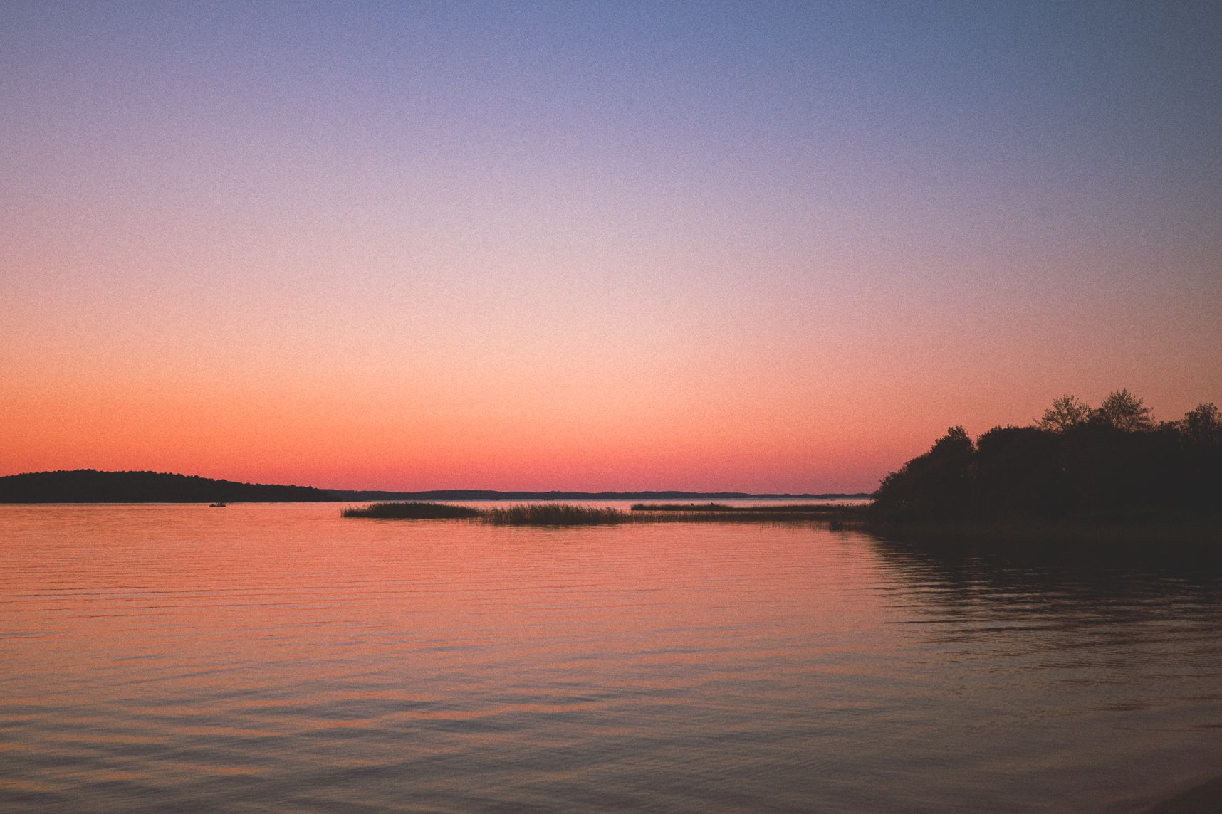 Le lac est calme et le soleil donne ses dernières lueurs. Le ciel est un dégradé orange qui reflète sur l'eau. Au milieu des roseaux qui sortent tout juste de l'eau