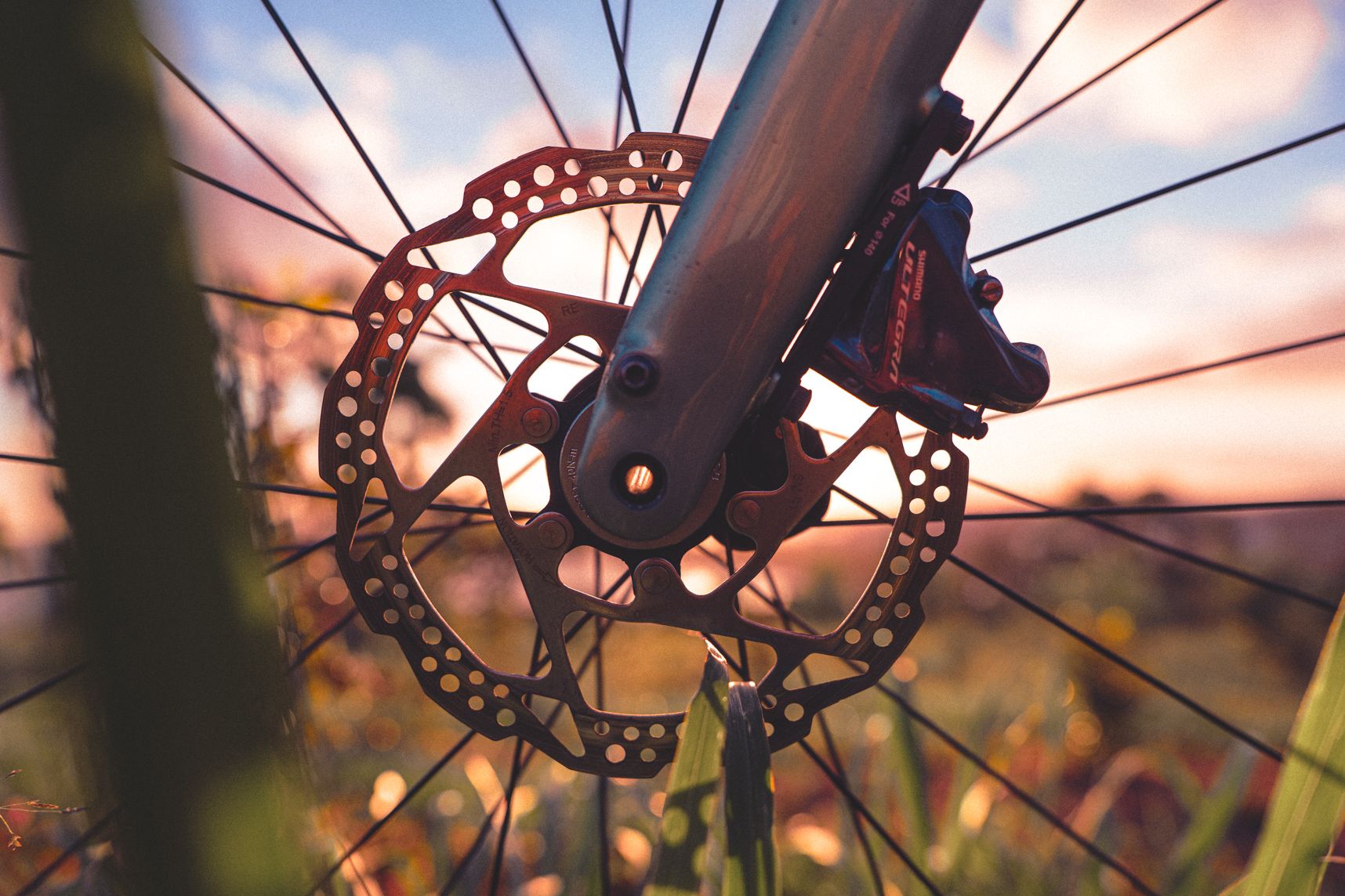 Gros plan sur le frein à disque d'un vélo. La roue est dans les hautes herbes. Derrière le coucher de soleil et les nuages