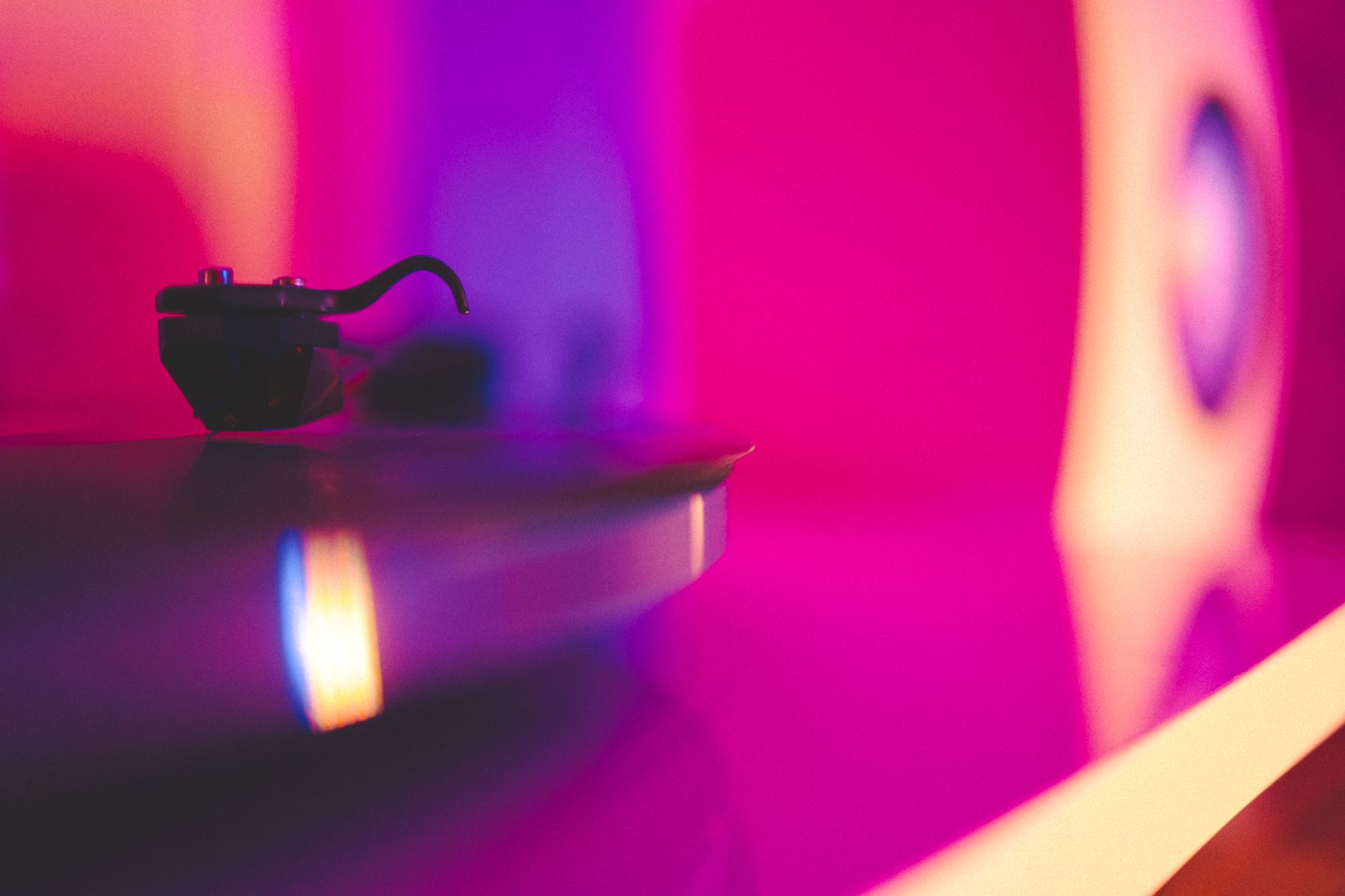 Gros plan sur la tête de lecture d'une platine vinyle. L'ambiance est rose. Derrière dans le flou une enceinte
