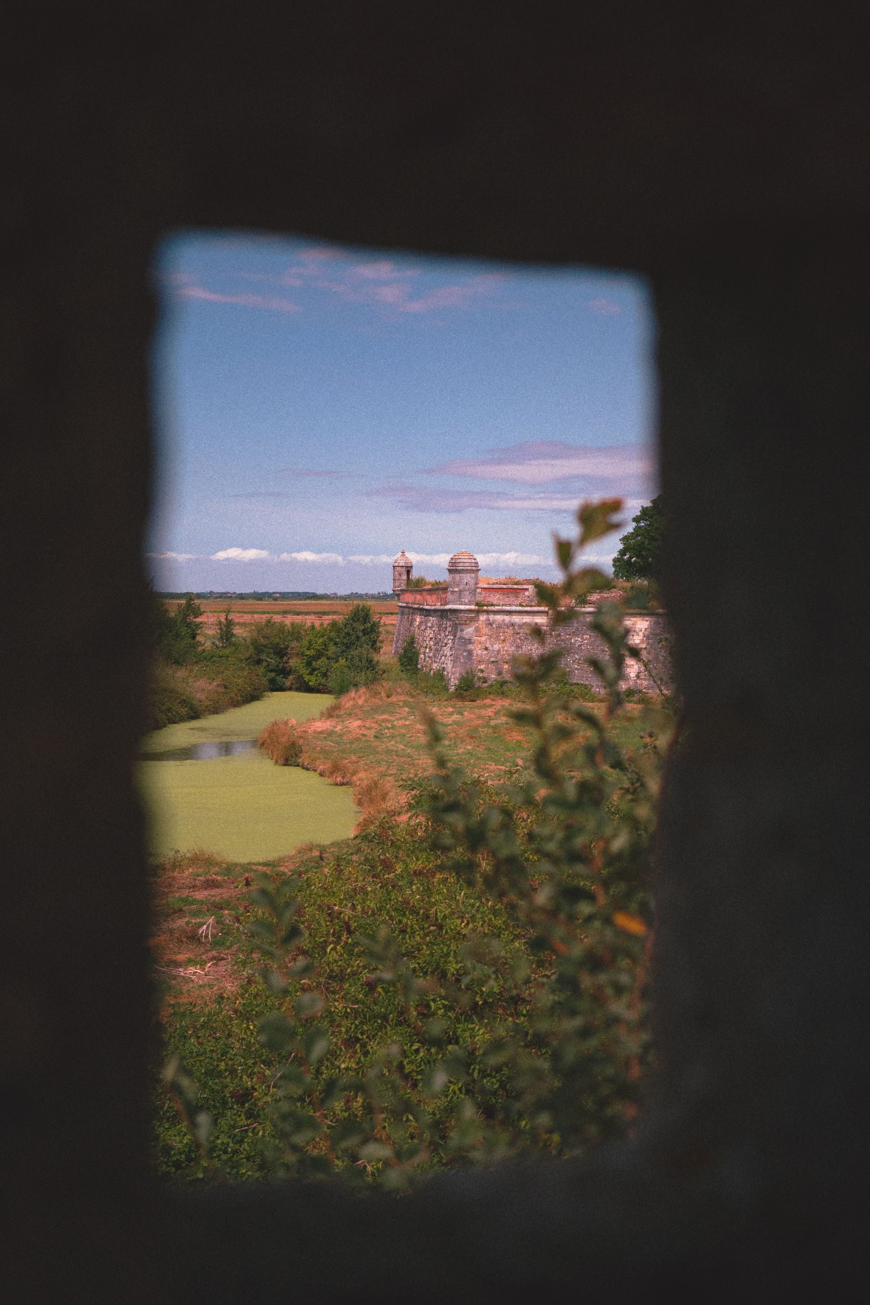 Vue à travers une petite fenêtre d'une tour. On y voit les remparts de Brouage et sa douve végétalisée 