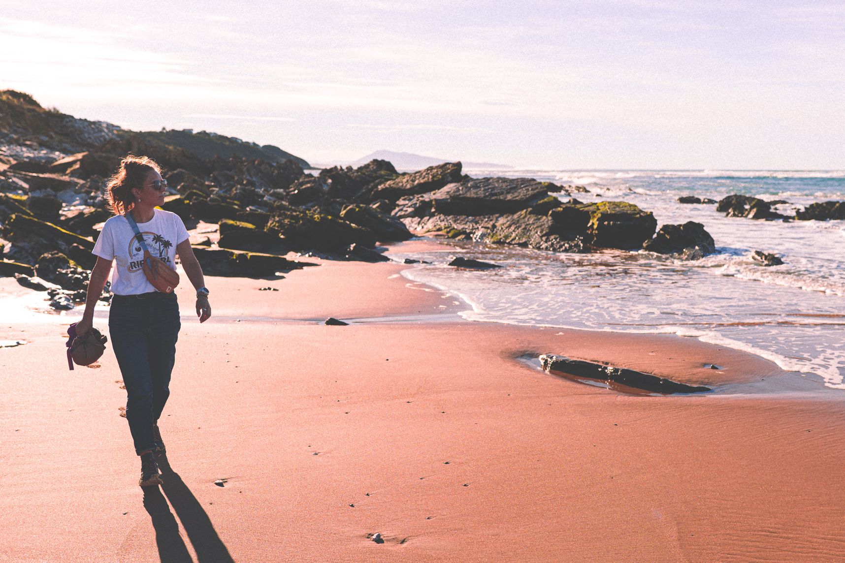 Une femme marchant de bonne allure sur la plage. Sur la droite la mer. Derrière les rochers et au loin la montagne. Le soleil est dans le dos.