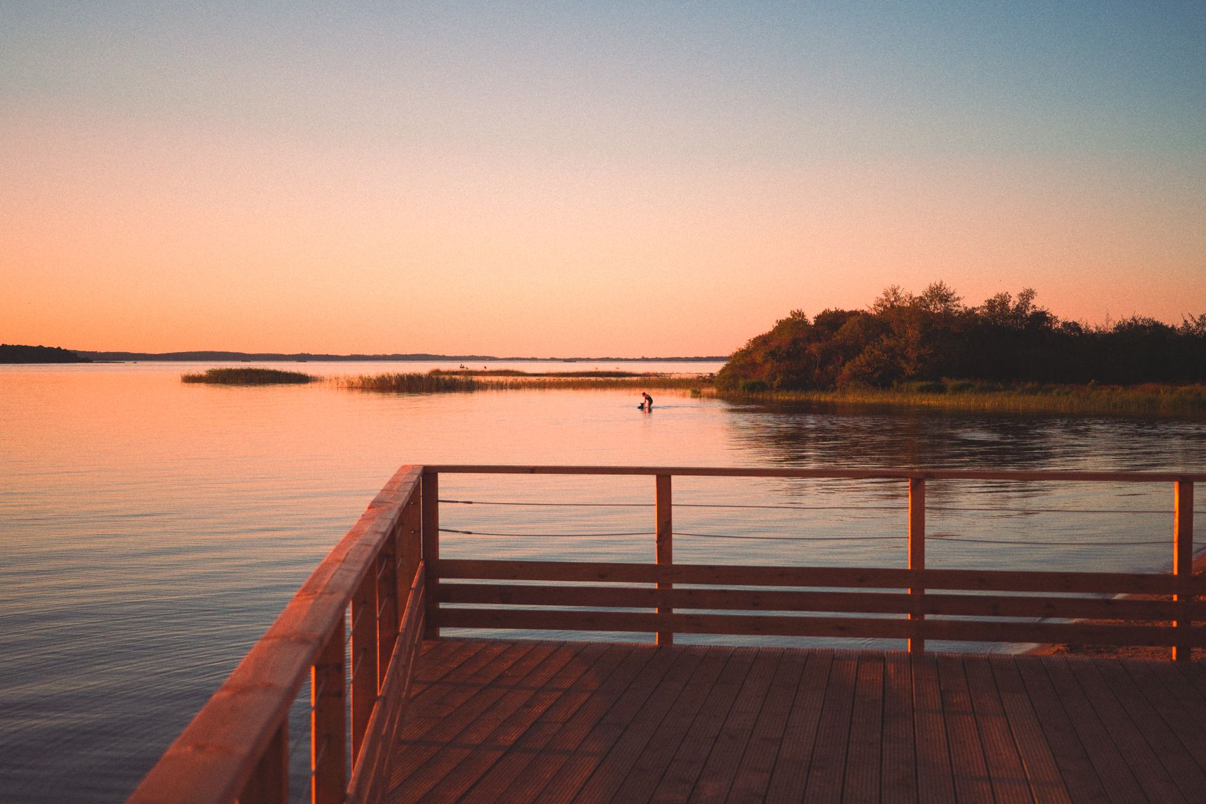 L'eau de l'étang de Biscarrosse est clame, c'est le coucher de soleil. Le ponton a des tons orangés. Dans l'eau une femme joue avec son chien