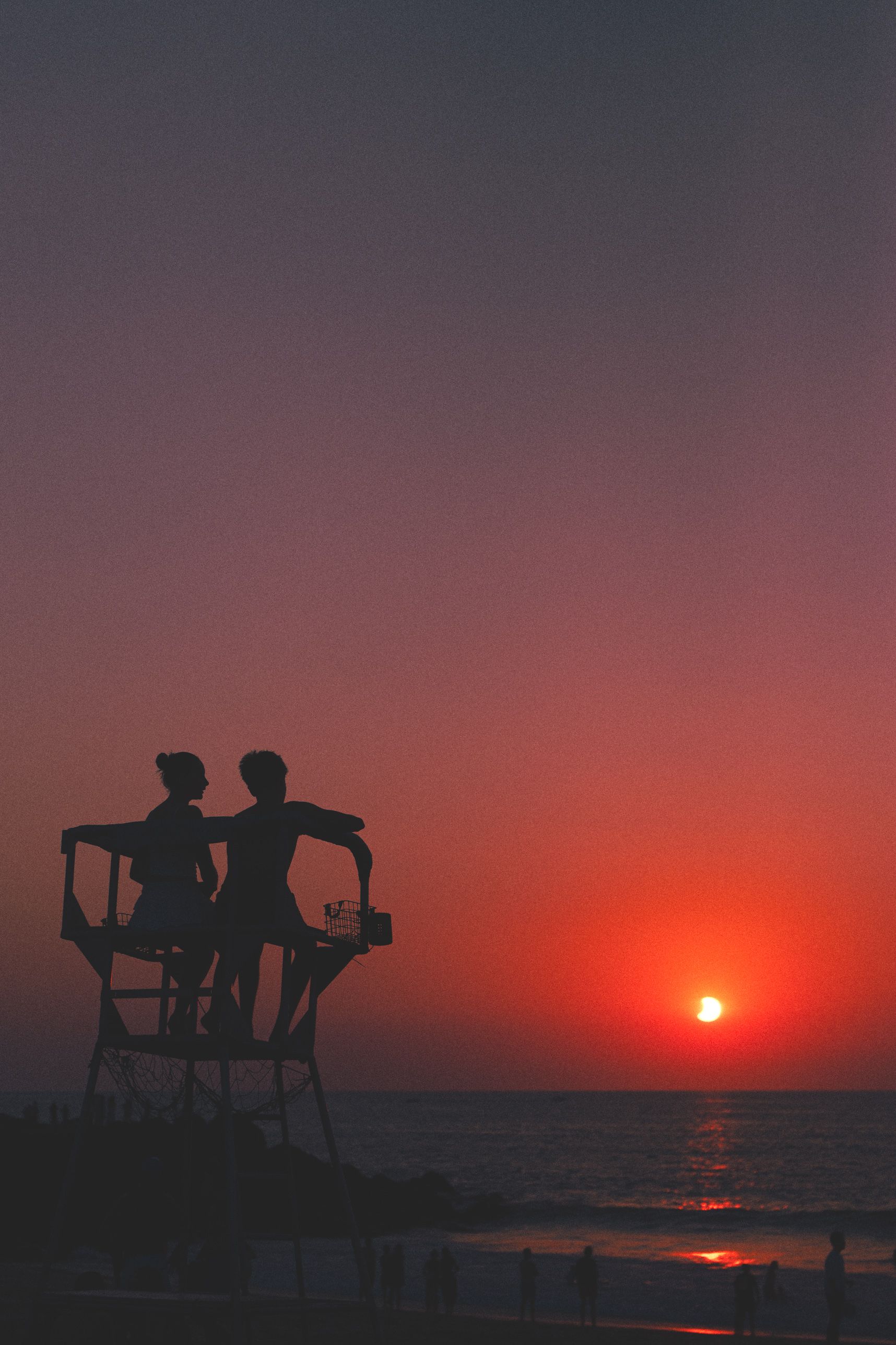 Deux personnes en contre-jour, debout sur une plateforme de poste de secours en bord de plage, regardent le soleil se coucher à l'horizon dans un ciel dégradé du violet au rouge. Quelques promeneurs marchent au bord de l'eau.