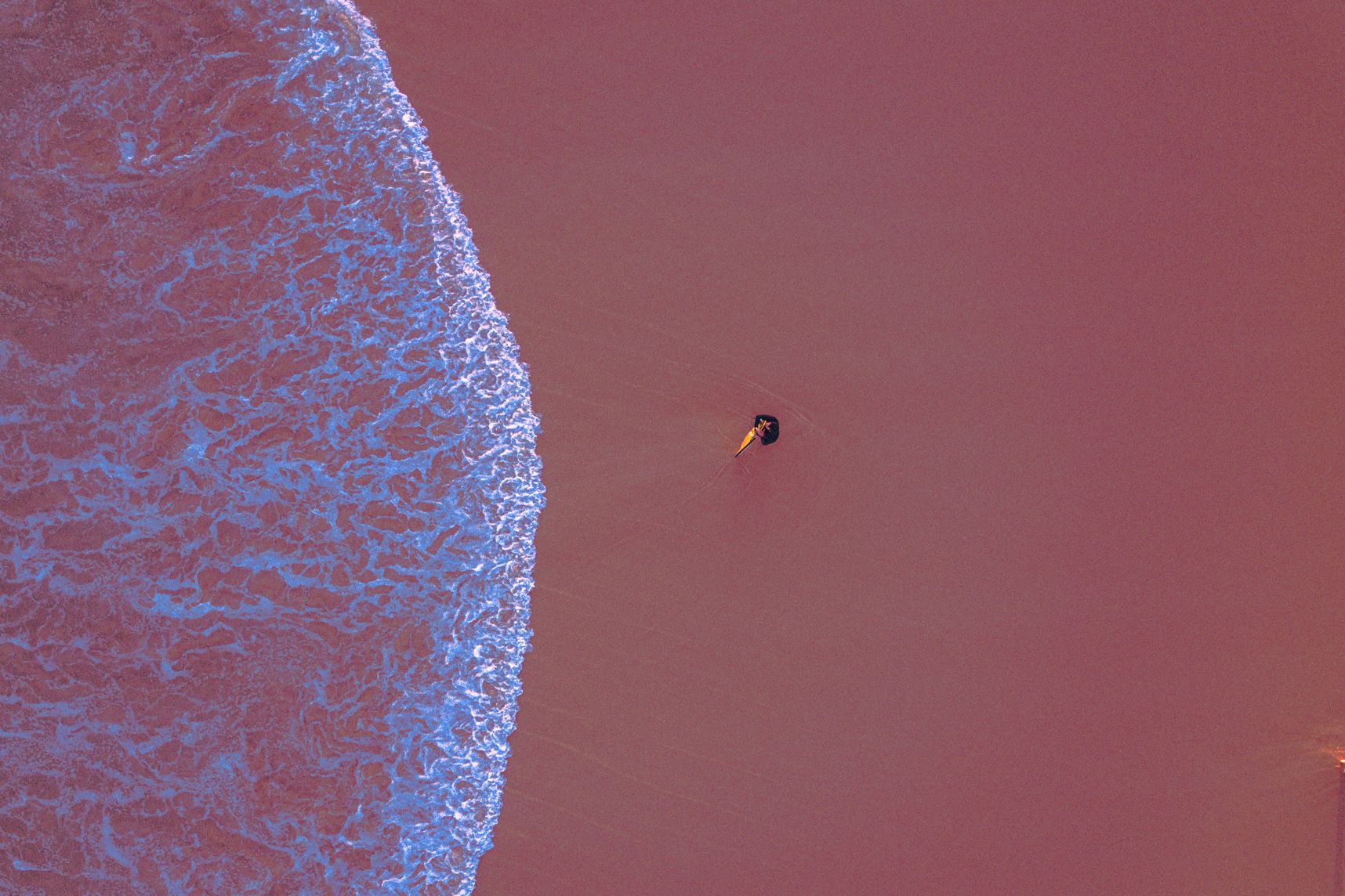 Vue de drone vers le sol, un surfeur tien sa planche dans ses bras et regarde l'horizon alors qu'une vague arrive.  La form et la couleur blanchâtre de la vague fait penser à une méduse