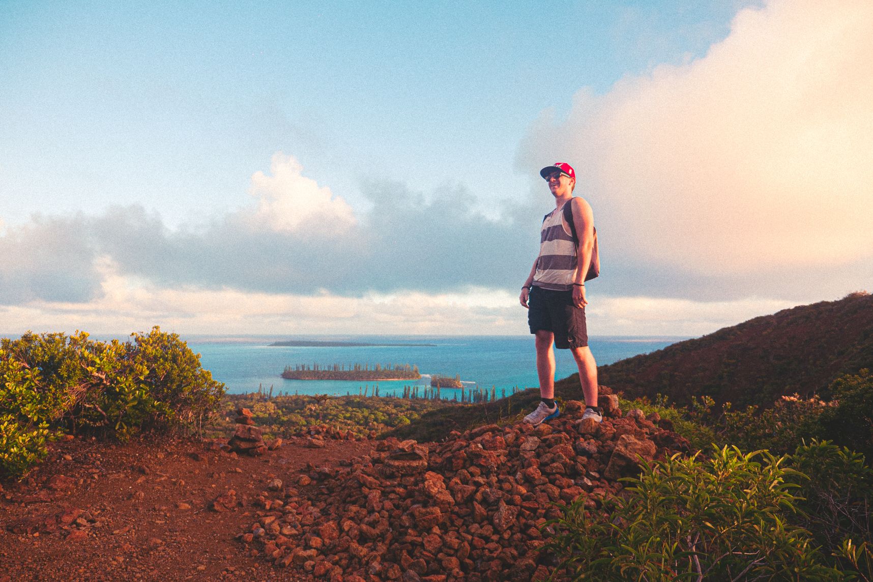 Arrivé en haut de la randonnée sur l'ile des Pins, le paysage est magnifique. Un homme debout sur un tas de cailloux à le sourire