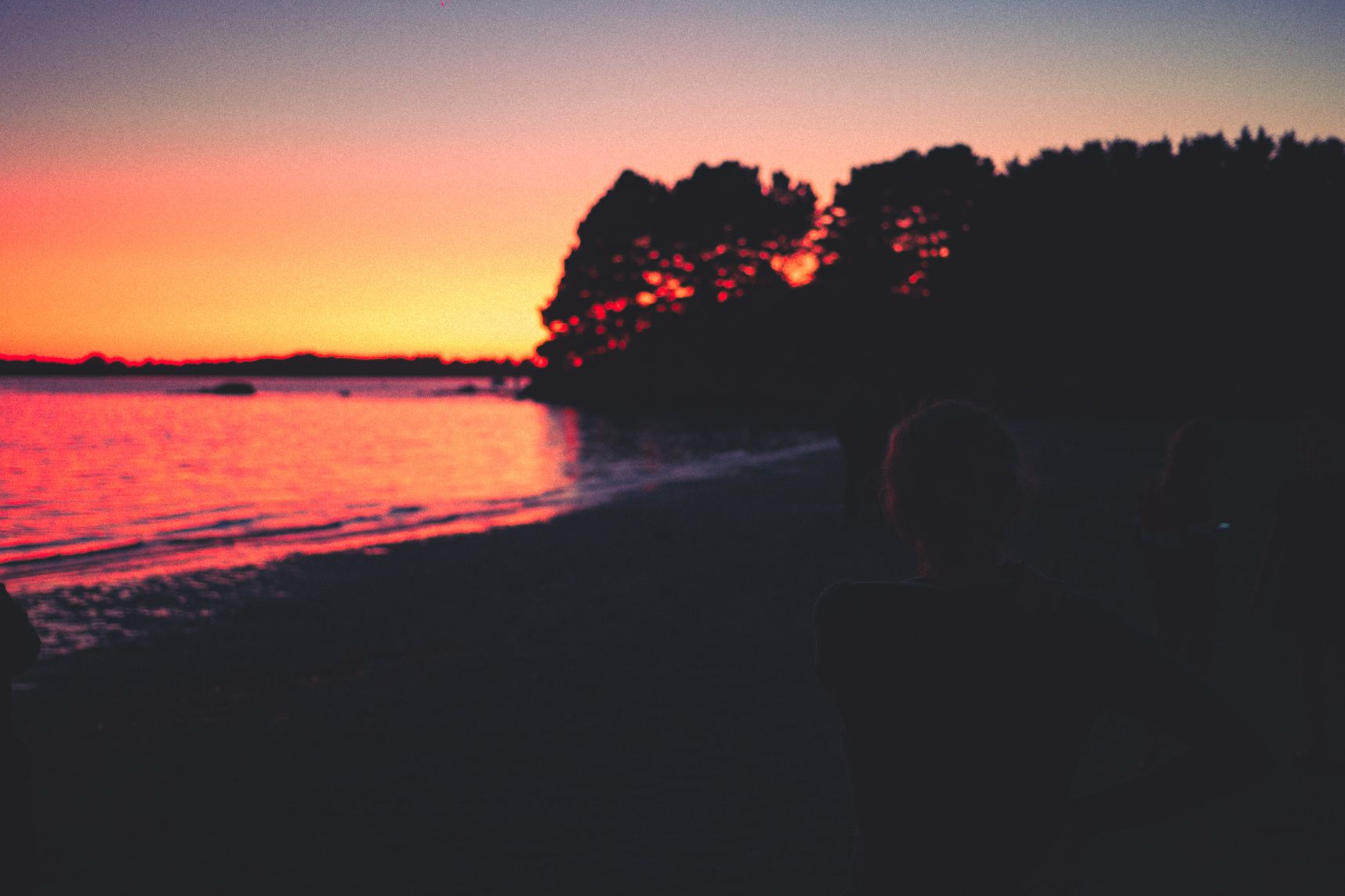 Le soleil se couche derrière les arbres, sa lueur orangée reflète sur l'eau, les gens admirent le paysage