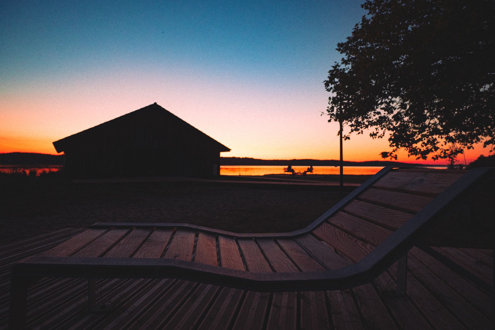 Le soleil est couché sur l'étang de Biscarrosse et de Parentis, on y voit encore le ciel orangé. En premier plan une chaise longue parfaite pour admirer la nature. Plus loin une cabane dans l'ombre et un groupe d'amis à l'apéro