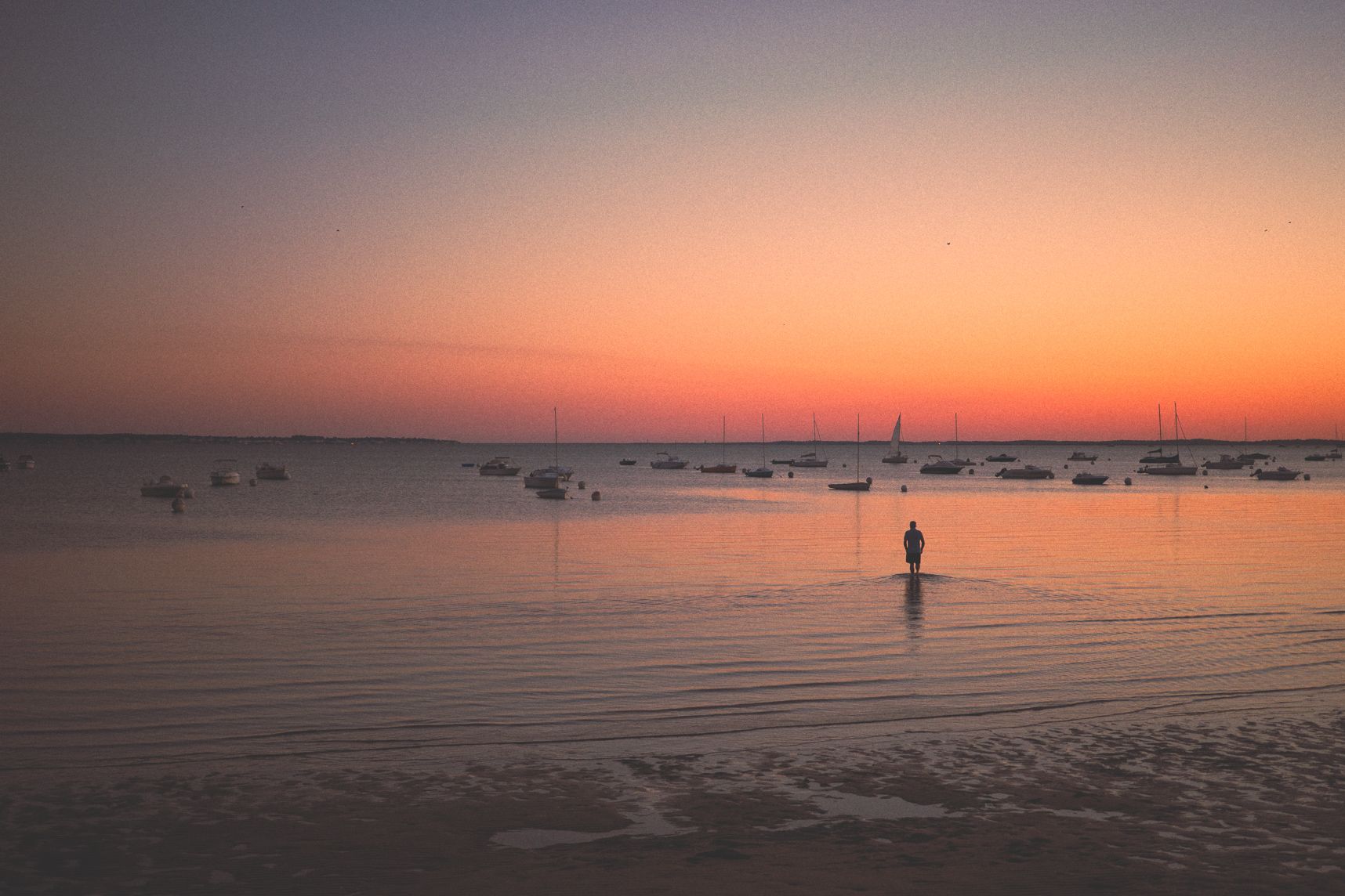 Au coucher de soleil, l'eau du bassin d'Arcachon est calme. Une personne avance vers son bateau les pieds dans l'eau