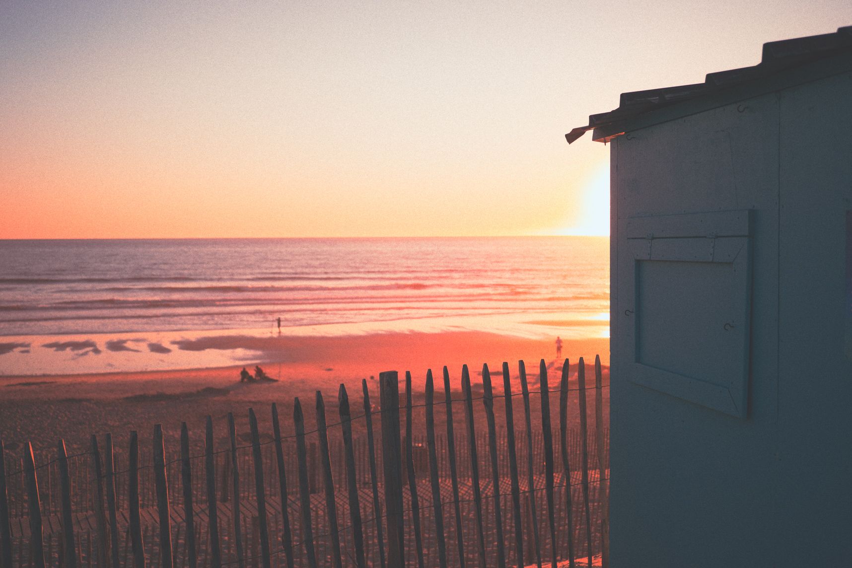 La cabane bleu en haut de la dune cache partiellement le coucher de soleil. Sur la plage les gens admirent le paysage. Il y a quelques vagues